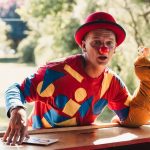 A clown wearing a colorful costume entertains with a puppet outdoors on a sunny day.